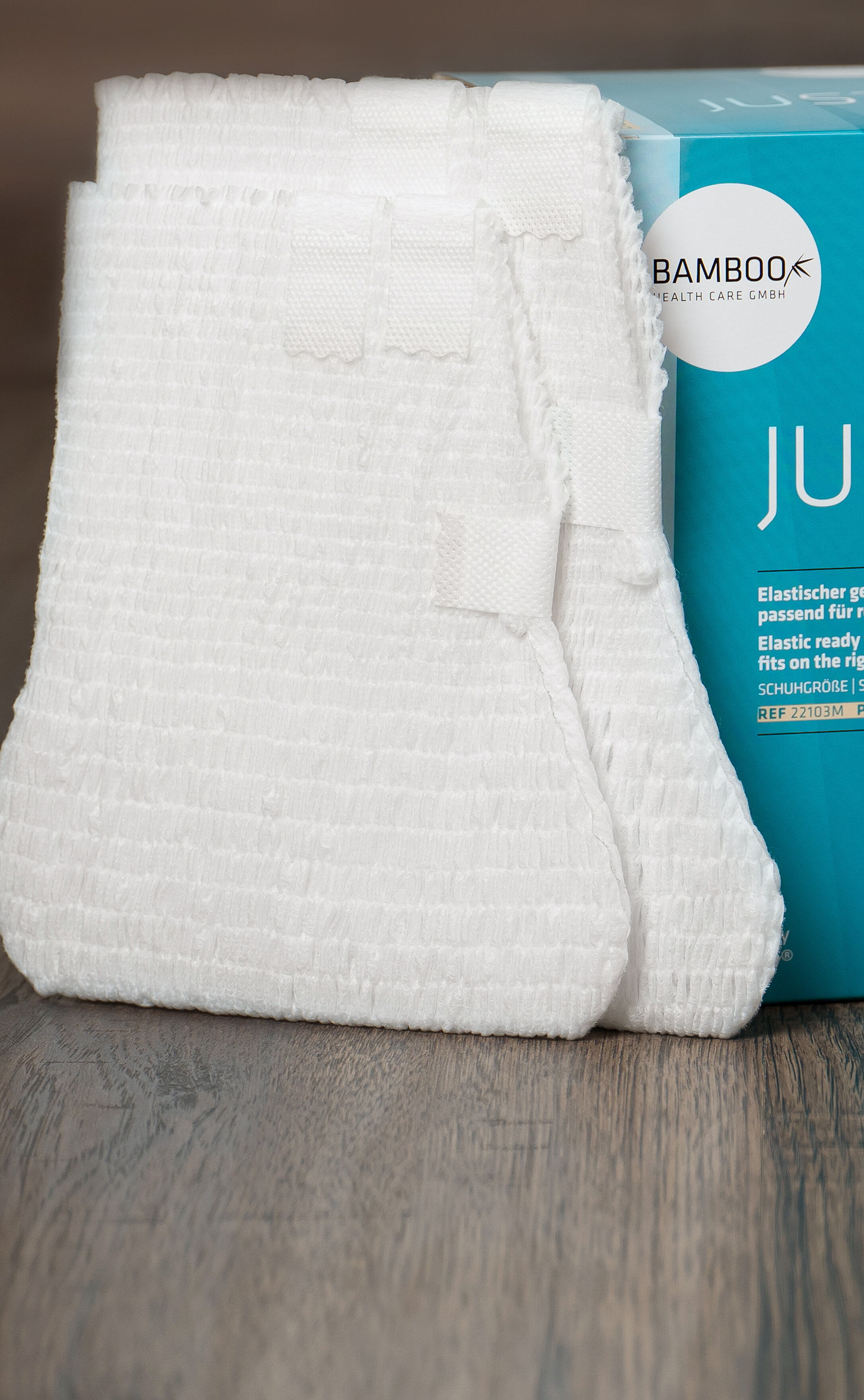 Zwei unterschiedliche Verpackungsgrößen von JustSox® stehend mit zwei Fußverbänden im Vordergrund auf einem dunklen Holzboden. | © Bamboo Health Care GmbH