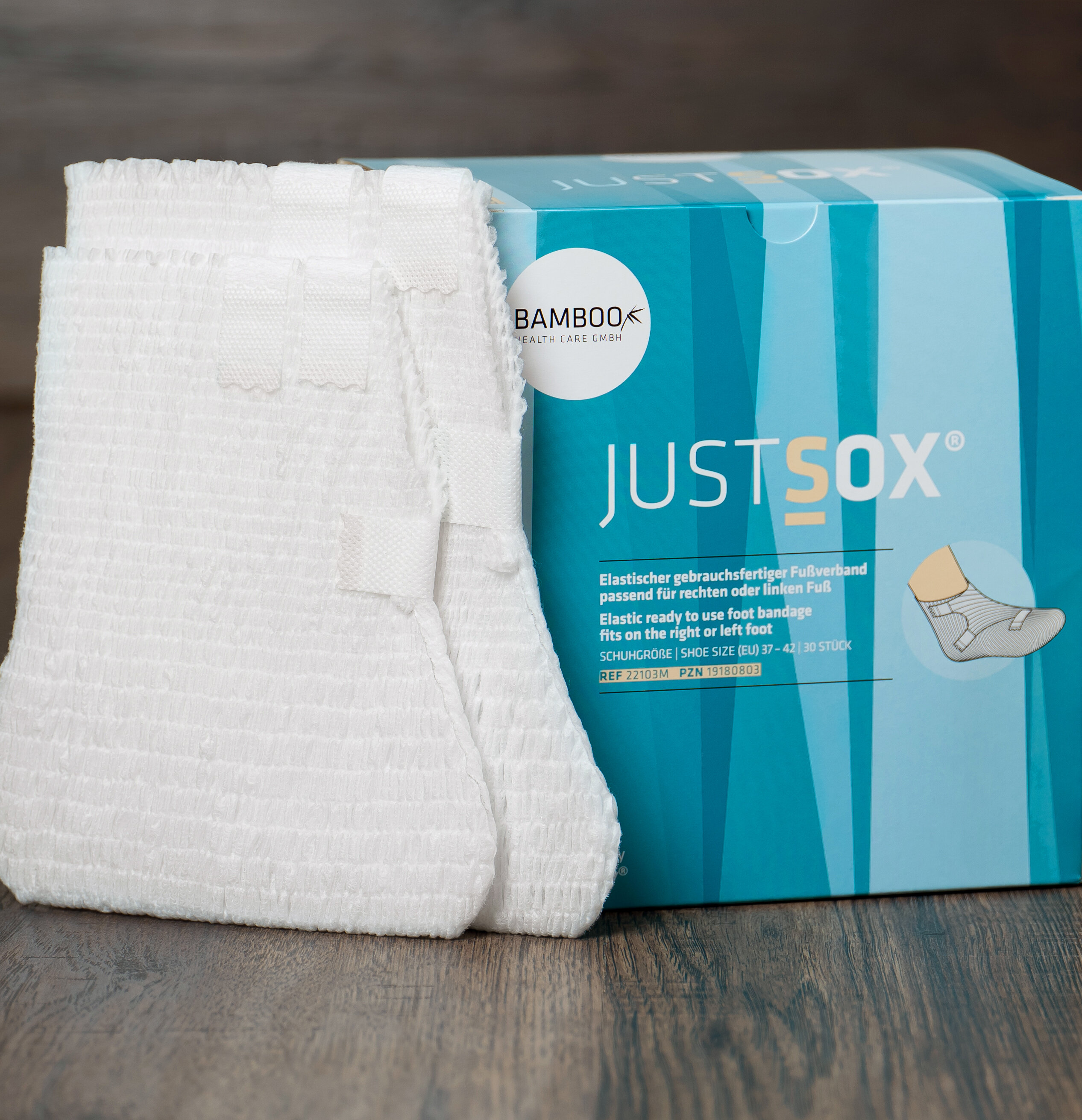 Eine Verpackung von JustSox® stehend mit zwei Fußverbänden im Vordergrund auf einem dunklen Holzboden.